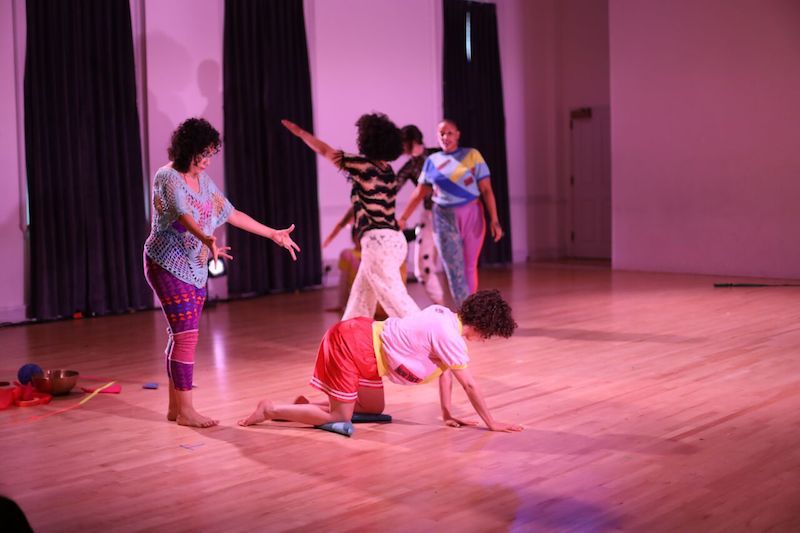 Dancers stand in colorful clothes. One woman crawls.